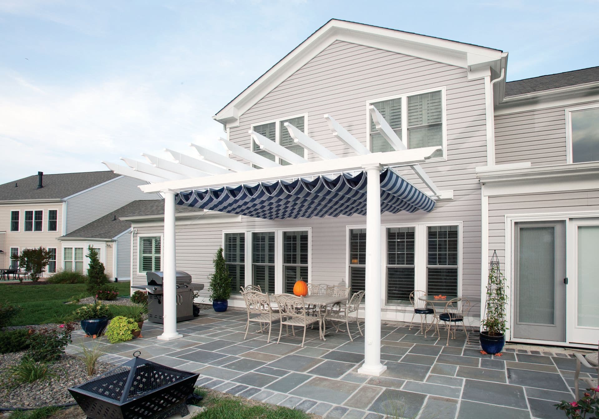 Patio with white pergola and blue awning attached to a gray house. Features a dining set, a pumpkin centerpiece, and a landscaped garden. Calm, inviting vibe.