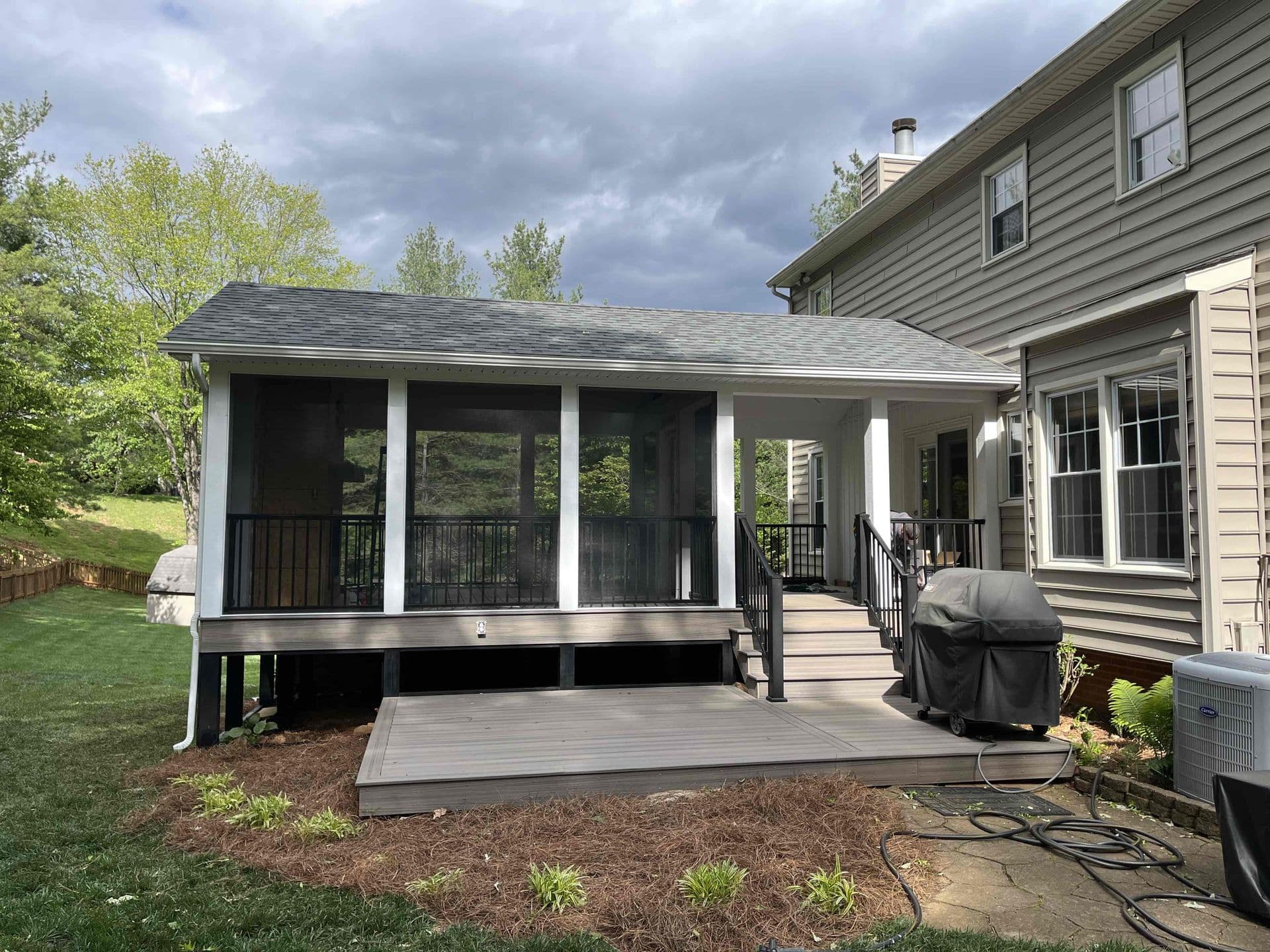 Suburban house with a screened porch and deck featuring a grill. Overcast sky and surrounding green lawn suggest a calm, residential setting.